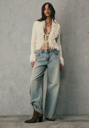 Cardigan corto, cremoso e morbido con chiusura a legatura e maniche a coste, abbinato a jeans a vita alta in denim chiaro e stivaletti marroni con tacco.