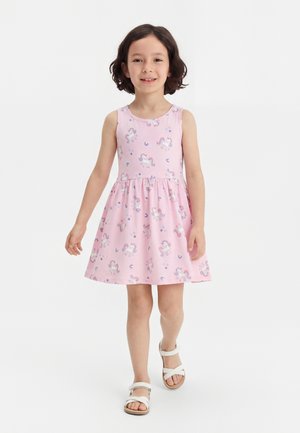 Νεαρό κορίτσι περπατάει με ροζ αμάνικο φόρεμα με σχέδια μονόκερων και λευκά σανδάλια σε απλό λευκό φόντο.