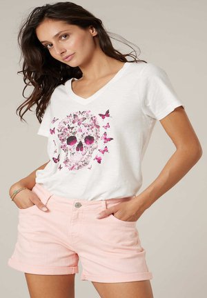 Femme portant un T-shirt blanc à col en V avec un motif de papillon rose en forme de crâne et un short rose pâle à ourlet roulé, mains dans les poches, arrière-plan neutre.