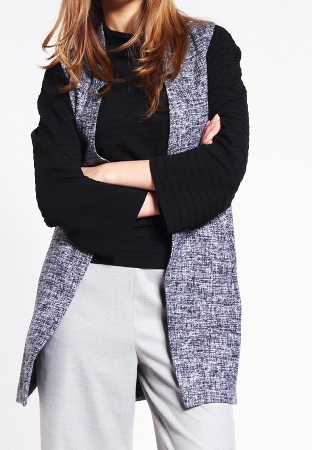 Longue veste grise avec manches texturées noires, portée par-dessus un haut côtelé noir. La tenue comprend un pantalon gris clair. Aucun motif ni accent visible.