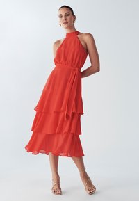 Rode chiffonjurk met een halternek, gelaagde ruches en een gestrikte taille. Gecombineerd met gouden sandalen met bandjes. Zachte textuur en vloeiend ontwerp.