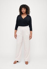 Top avvolgente nero a maniche lunghe abbinato a pantaloni ampi beige chiaro a vita alta, con dettagli di cintura e sottili pieghe.