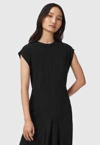 Černé šaty s krátkými rukávky, hladkou texturou, kulatým výstřihem a ostrými švy, které zvýrazňují splývavou siluetu.