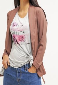 Blazer marrone, leggero, con apertura frontale e maniche lunghe, abbinato a una maglietta grigia con grafiche floreali e un dettaglio annodato in vita.