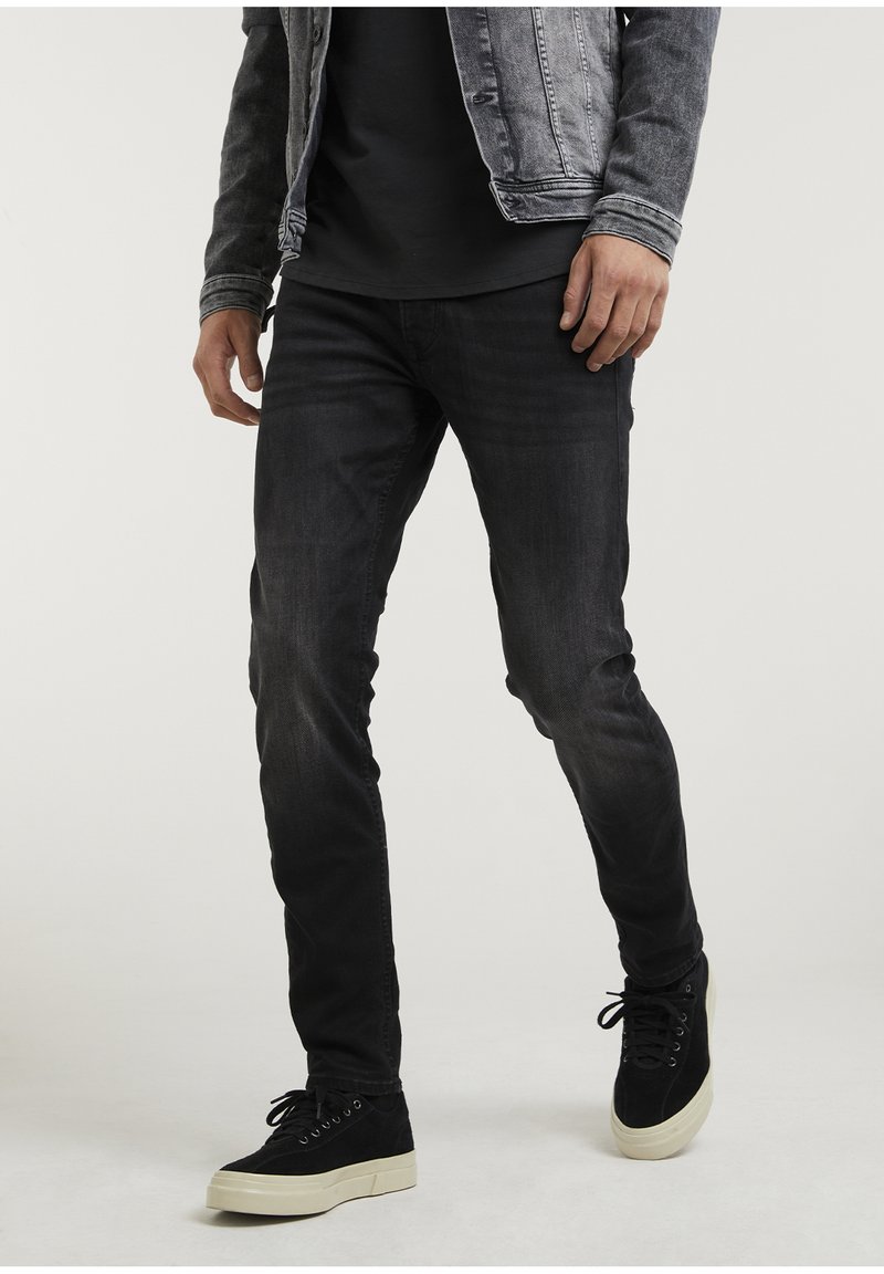 Man in zwarte slim-fit jeans, zwarte sneakers met witte zolen, een zwart t-shirt en een grijze spijkerjas, staand voor een effen achtergrond.