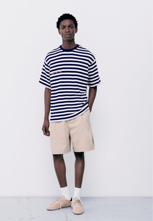 Jeune homme debout portant un t-shirt rayé bleu marine et blanc, un short beige, des chaussettes blanches et des baskets beiges sur un fond clair uni.