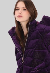 Fioletowa kurtka puchowa o błyszczącej teksturze, pikowanym wzorze, z zamkiem błyskawicznym oraz kapturem obszytym futerkiem. Charakteryzuje się dopasowanym krojem i ozdobnymi guzikami.