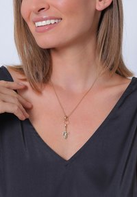 Collar de tono dorado con un pequeño colgante en forma de figura, colgando de una delicada cadena. Lleva un top oscuro con escote en V.