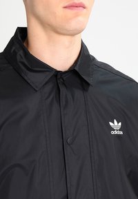 Svart vindjacka med krage, tryckknappar och en vit Adidas-logotyp på vänster bröst. Tillverkad av ett lätt och mjukt material.