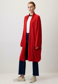 Červený dlhý kardigán vyrobený z mäkkého, textúrovaného materiálu. Má otvorený predný diel, dlhé rukávy a predné vrecká. Skombinovaný s tmavými džínami a svetlými topánkami.