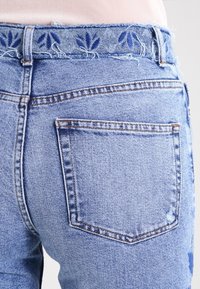 Světle modré džínové kalhoty s třepícím se pasem a jemnou květinovou výšivkou. Obsahují dvě zadní kapsy a mírně vybledlé oblasti s opotřebením.