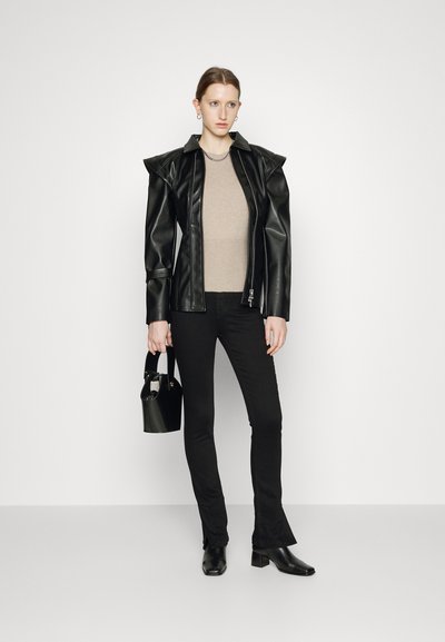 Jaqueta de couro sintético preta com acentos nos ombros, blusa justa bege, calças pretas e botas de salto. Modelo segurando uma pequena bolsa preta.