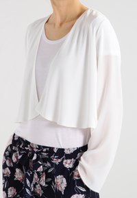 Bílý zkrácený cardigan s volnými dlouhými rukávy, vyrobený z lehkého materiálu, vrstvený design, spárovaný s květovanými kalhotami.