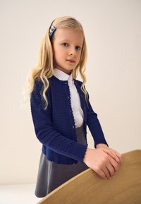 Marineblauwe gebreide cardigan met structuurpatroon en gegolfde zoom, gedragen over een wit overhemd met kraag. Model met lang blond haar poseert bij een houten tafel.