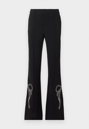 Kobieta stoi z dłońmi w kieszeniach, ubrana w srebrny bezrękawnik, czarne rozszerzane spodnie z białą haftem i czarne spiczaste buty.