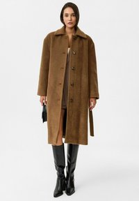 Bruine wollen mix mantel met een kraag, knopen vooraan en twee zakken. Gedragen over een beige mini-jurk en gecombineerd met zwarte laarzen tot de knie.