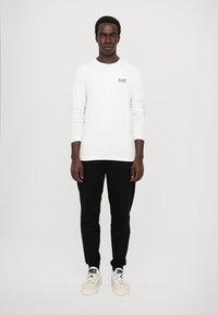 Langermet hvit T-skjorte med EA7-logo, kombinert med svarte bukser og hvite sneakers på en modell foran en enkel bakgrunn.