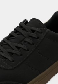 Pantofii sport negri au un superior dintr-un material textil neted, sistem de prindere cu sireturi, detalii de cusătură subtile și o talpă din cauciuc maro contrastantă.