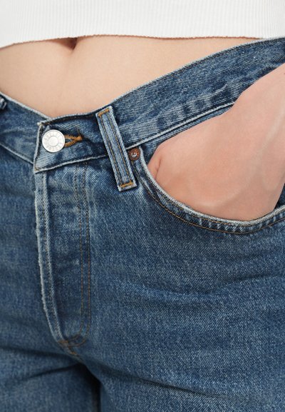Jean en denim bleu avec une taille haute, présentant des coutures accentuées, une fermeture à bouton métallique et une poche avant. Texture lisse.