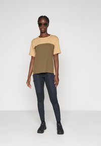 Kvinne med solbriller, en brun og oliven kortermet skjorte, svarte og marineblå leggings med lommer, og svarte treningssko, stående mot en hvit vegg.