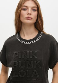 Black short-sleeve t-shirt featuring textured "PINK PUNK LOLA" print and a beaded neckline accent with a smooth cotton finish.
