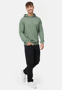 Grüner Kapuzenpullover mit verstellbarem Kapuzenzug und gestreiften Kordeln, kombiniert mit schwarzen Hosen und weißen Sneakers. Das Modell steht vor einem neutralen Hintergrund.