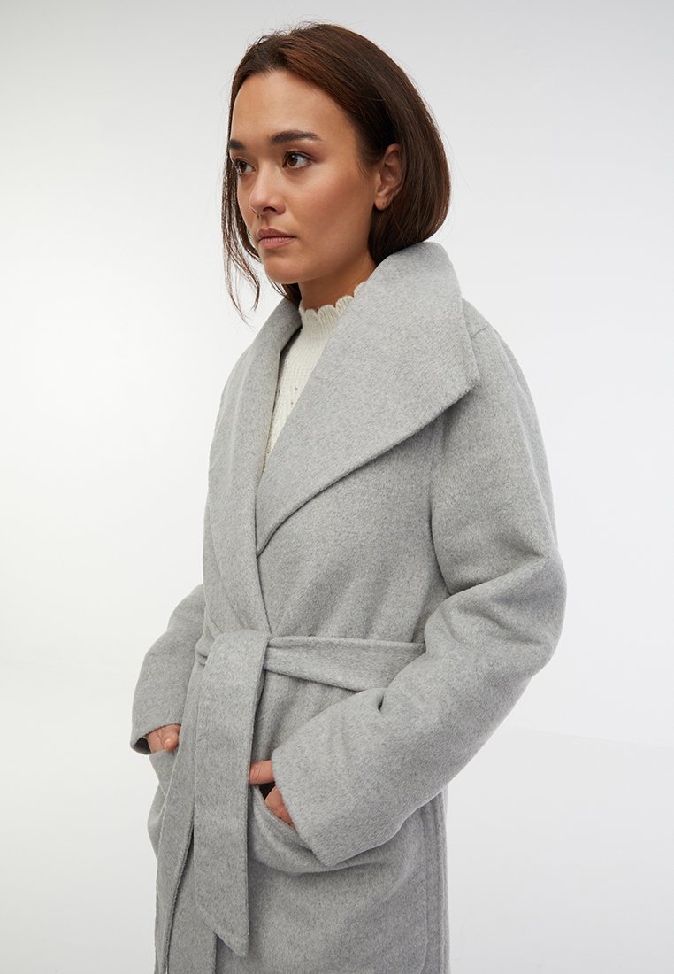 Kobieta z brązowymi włosami, ubrana w jasnoszary wełniany płaszcz z paskiem, na białym swetrze, stojąca z rękami w kieszeniach płaszcza, spoglądająca w lewo.
