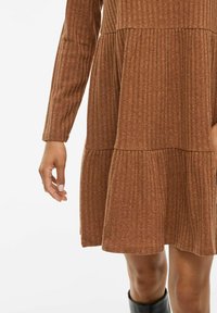 Braunes geripptes Kleid mit langen Ärmeln, gestuften Design und strukturiertem Stoff. Sichtbare Hand mit manikürten Nägeln und schwarzem Schuhwerk.