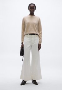 Beżowy wełniany sweter, białe spodnie o szerokich nogawkach, czarny pasek i czarna torebka. Gładka tekstura, prosty fason i prosta sylwetka.