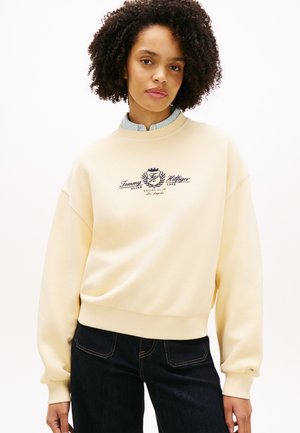 Mlada ženska z kodrastimi lasmi, oblečena v kremno Tommy Hilfiger majico z dolgimi rokavi in temne kavbojke, stoji pred enobarvnim belim ozadjem.