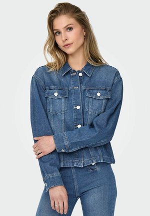 Young woman with long blonde hair wears blue denim jacket and matching jeans, standing with arms crossed against plain background.
