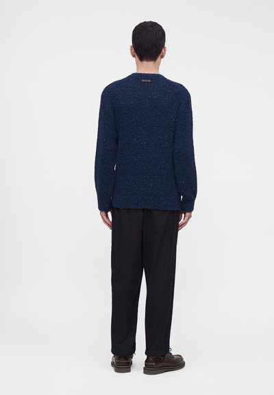 Homme de dos portant un pull bleu marine texturé, un pantalon noir ample et des chaussures marron foncé à lacets sur un fond clair uni.