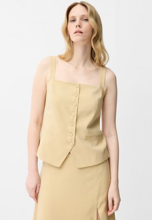 Donna con capelli biondo chiaro che indossa un top beige senza maniche abbottonato e gonna coordinata su sfondo bianco.