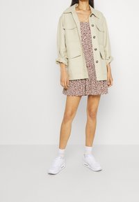 Vaaleanbeige takki, jossa on napit edessä ja useita taskuja, ruskean ja valkoisen leopardi-printtisen mekon päällä, yhdistettynä valkoisiin tennareihin ja sukkiin.