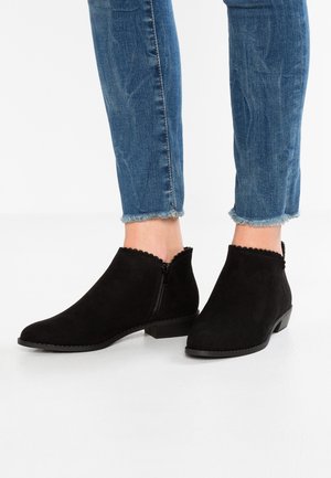 Fødder iført sorte ruskind ankelstøvler med bølgede kanter og sidesyninger, kombineret med blå jeans med frynsete kanter.