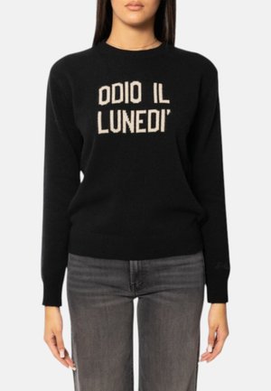 Donna con indosso un maglione nero con testo bianco che recita "ODIO IL LUNEDÌ" e jeans grigi scoloriti, in piedi davanti a uno sfondo semplice.