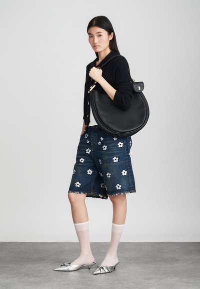 Femme jeune portant un cardigan navy, un short en jean brodé, des chaussettes blanches transparentes, des talons en argent, tenant un grand sac à main en cuir noir sur l'épaule.