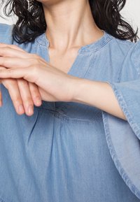 Osoba s kudrnatými vlasy, oblečená v denimovém topu, je zobrazena se zaměřením na jejich ruce, které jemně spočívají spolu blízko výstřihu.