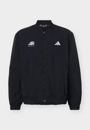 Νέος άνδρας με μαύρο μπουφάν Adidas με φερμουάρ και λογότυπα, και κόκκινα αθλητικά σορτς, στέκεται μπροστά σε καθαρό φωτεινό φόντο.
