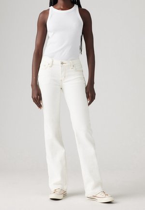 Osoba w białej bluzce bez rękawów, wysokich, jasnobeżowych dżinsach i białych trampkach, stojąca na jasnym, jednolitym tle.