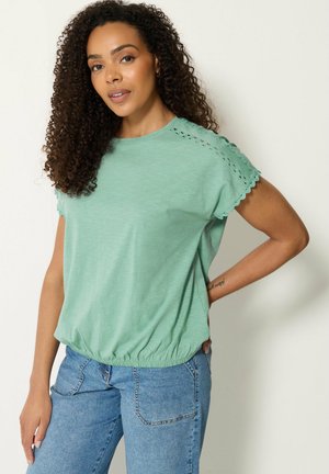 Vrouw met krullend haar, gekleed in een muntgroen kortemouwtopje met kantdetails op de schouders en blauwe jeans, poserend met één hand op de heup.