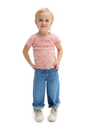 Małe dziecko o blond włosach, ubrane w różową koszulkę w serduszka, luźne niebieskie dżinsy z podwiniętymi nogawkami i beżowe trampki, stojące z rękami na biodrach.