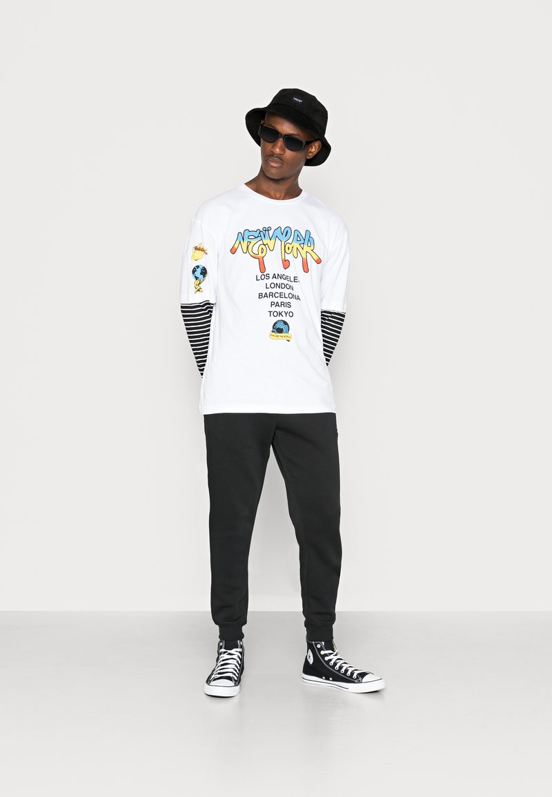 Hvit langermet t-skjorte med fargerik "New York" grafikk; svart og hvit stripete mansjetter. Kombinert med svarte bukser og svarte høye sneakers.