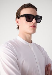Black square sunglasses with dark lenses and gold accent logo on the arm, paired with a white collared shirt. Smooth materials and clean lines.