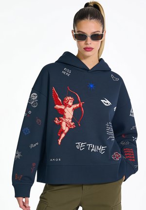 Femme portant un sweat à capuche bleu marine avec un motif de cupidon rouge et un texte sur le thème de l'amour, associé à des lunettes de soleil et un pantalon vert olive.