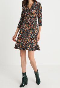 Kwiatowa sukienka w stylu wrap z dekoltem w serek, rękawami 3/4, na ciemnym tle, z wcięciem w talii i rozkloszowanym dołem, w zestawieniu z zielonymi botkami na obcasie.