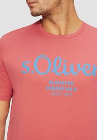 Lyhythihainen t-paita korallinpunaisena, jossa pyöreä kaula-aukko. Edessä sinisellä painettuna teksti: "s. Oliver ELEVATED ESSENTIALS SINCE 1969".