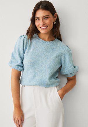 Kvinde smiler, iført lyseblå trøje med pufærmer og hvide bukser, stående foran en ensfarvet lysegrå væg.