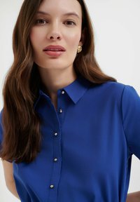 Camicia azzurra con colletto, maniche corte e dettagli in punta argentata sul davanti. Tessuto liscio, leggero e design aderente.