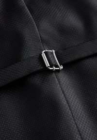 Gros plan sur un tissu noir texturé avec une boucle métallique argentée qui sécurise une sangle noire assortie.